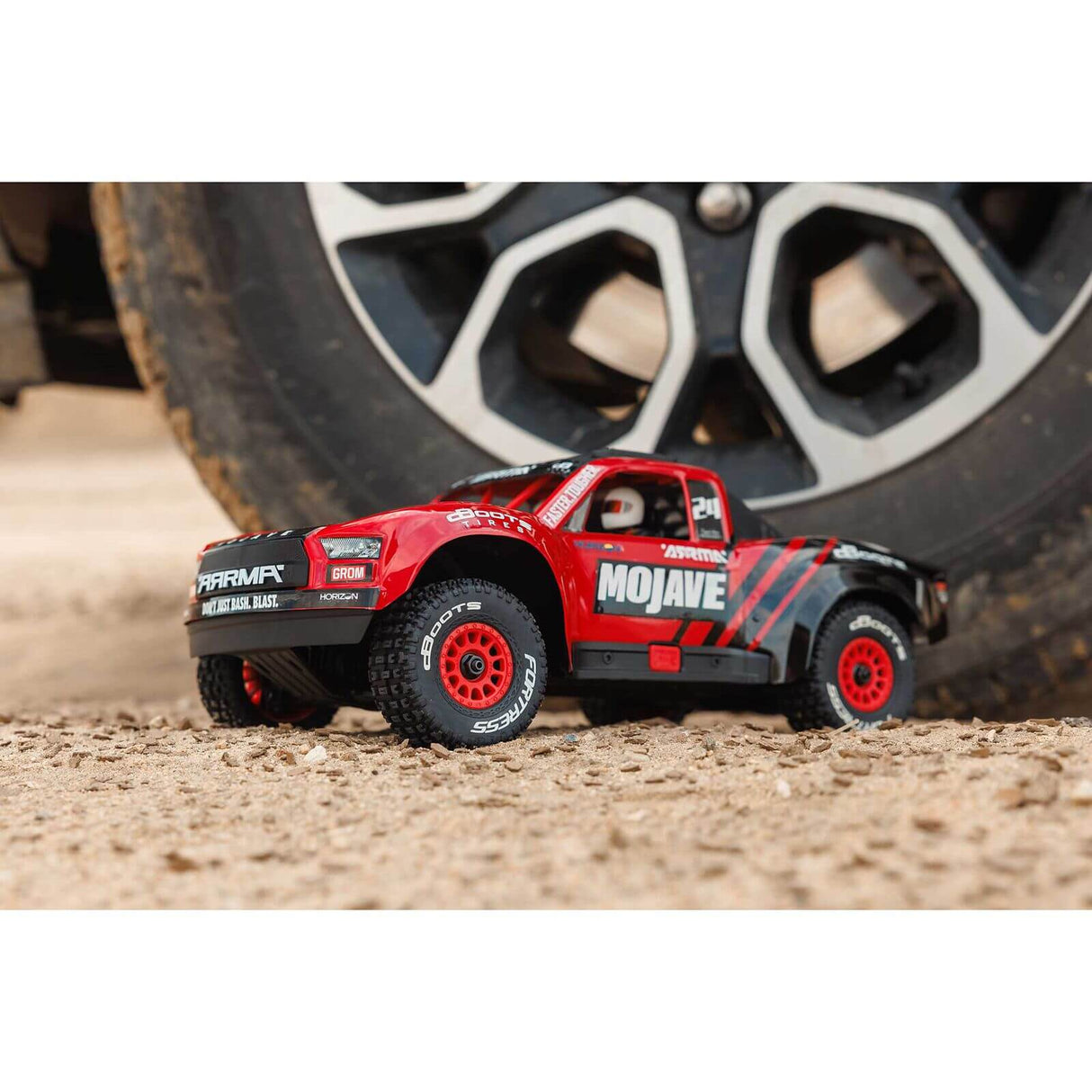 ARRMA Mojave Grom MEGA 380 Brushed 4X4 RC Desert Truck rot/schwarz neben großem Fahrzeugrad auf sandigem Untergrund